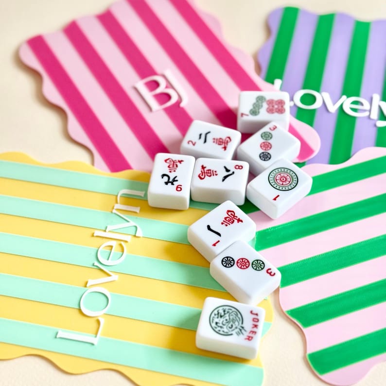 May include: A collection of white Mahjong tiles with red and black symbols, scattered on colorful striped placemats. The placemats are in shades of pink, green, yellow, and purple, with the words "Lovely" and "Over" visible.