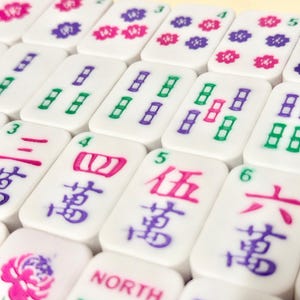 May include: White Mahjong tiles with colorful symbols and numbers. The tiles feature pink, purple, and green markings, including flower patterns and Chinese characters. The game pieces are arranged in a grid.