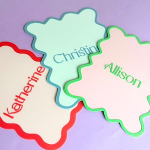 May include: Three name tags in a cloud shape with scalloped edges. The tags are in pastel colors: light pink, light blue, and off-white. The names "Allison", "Christin", and "Katherine" are printed on the tags in green, blue, and red.