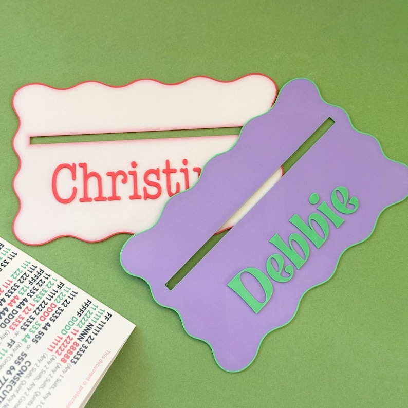 May include: Two personalized name tags with wavy edges. The top tag is white with red lettering that says "Christi". The bottom tag is purple with green lettering that says "Debbie".