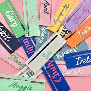 May include: A collection of colorful name tags in various hues, including pink, blue, green, and yellow. Each tag features a different name in a contrasting font. The tags are arranged on a pink background.