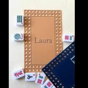 Pu&ograve; includere: Un sottopentola rettangolare beige con un bordo intrecciato e il nome "Laura" al centro. Diverse tessere di Mahjong bianche con simboli e numeri rossi e verdi sono sparse attorno al sottopentola, insieme a un tappetino blu.