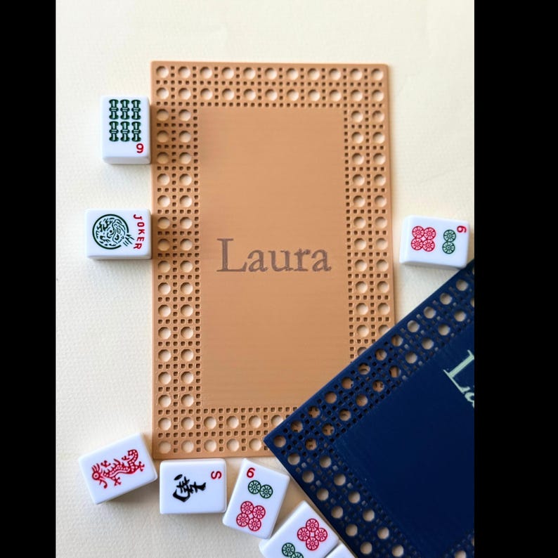Pu&ograve; includere: Un sottopentola rettangolare beige con un bordo intrecciato e il nome "Laura" al centro. Diverse tessere di Mahjong bianche con simboli e numeri rossi e verdi sono sparse attorno al sottopentola, insieme a un tappetino blu.