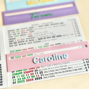 May include: A close-up of a pink card with the name "Caroline" in light blue letters. The card is on top of a sheet of paper with printed text and numbers. Other cards in blue and purple are in the background.