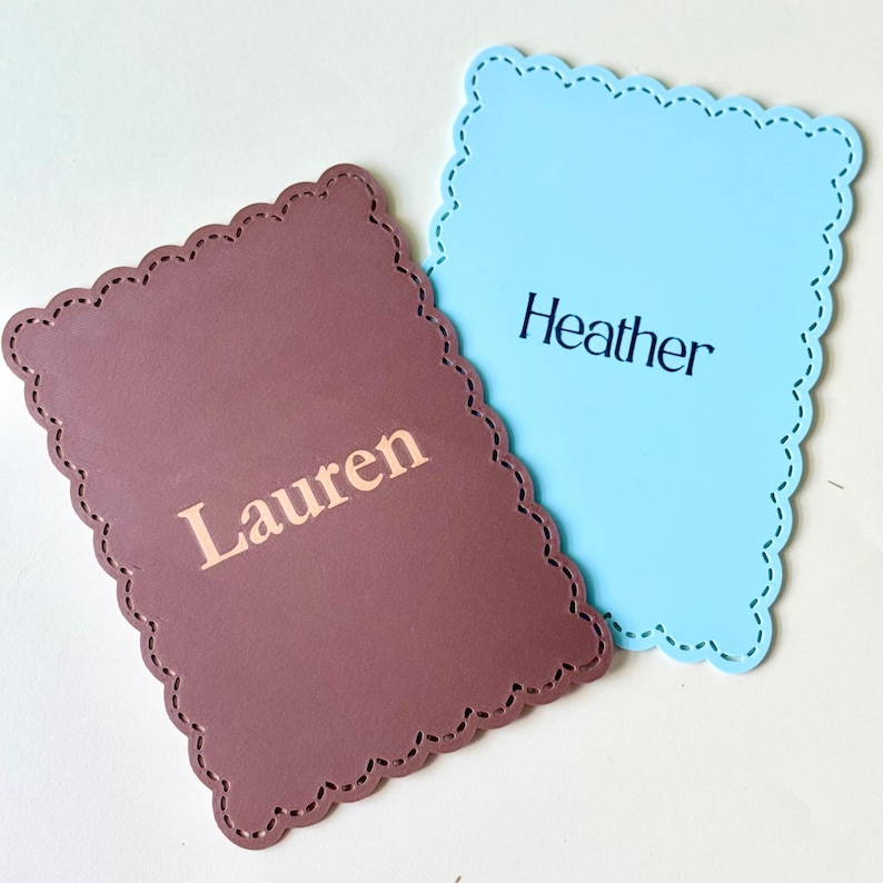 May include: Two name cards with a scalloped edge. One is brown with the name "Lauren" printed in a lighter colour. The other is light blue with "Heather" printed in black. Both cards have a decorative stitched border.
