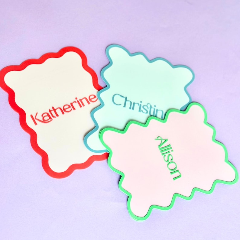 May include: Three decorative name tags with scalloped edges. One is cream-colored with a red border and the name "Katherine" in red. Another is light blue with a blue border and the name "Christine" in blue. The third is pink with a green border and the name "Allison" in green.
