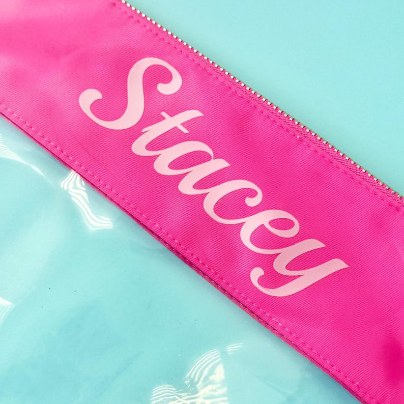 May include: A close-up of a pink cosmetic bag with the name "Stacey" printed in white script. The bag has a silver zip and is set against a light blue background. The bag appears to be made of a shiny, waterproof material.