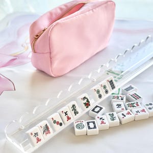 May include: A pink zippered cosmetic bag and a clear acrylic Mahjong tile rack with white tiles. The tiles display various symbols and numbers. The bag is made of a soft, quilted material.