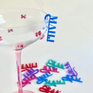 Può includere: Un bicchiere da cocktail trasparente con stelo rosa e decorazioni floreali, con un ciondolo blu che dice "MAUI". Diversi ciondoli colorati sono sparsi intorno al bicchiere.