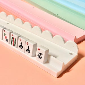May include: A white plastic mahjong tile rack with a scalloped top edge, holding several mahjong tiles featuring red, green, and black characters and symbols. Behind the white rack are additional racks in solid pastel pink and light blue colors.