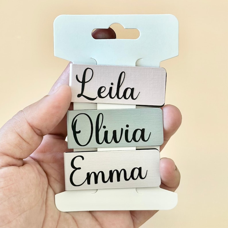May include: Three personalised metal name tags with the names Leila, Olivia, and Emma. The tags are stacked on top of each other and are displayed on a white card.