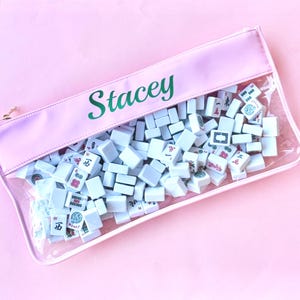 May include: A pink zippered pouch filled with white Mahjong tiles. The pouch has a clear front, allowing visibility of the tiles. The name "Stacey" is printed in green on the pink top. The background is a solid pink.