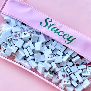 May include: A pink pouch containing white Mahjong tiles. The pouch has a clear front and a pink top with the name "Stacey" in green script. The tiles are white with various symbols and characters.