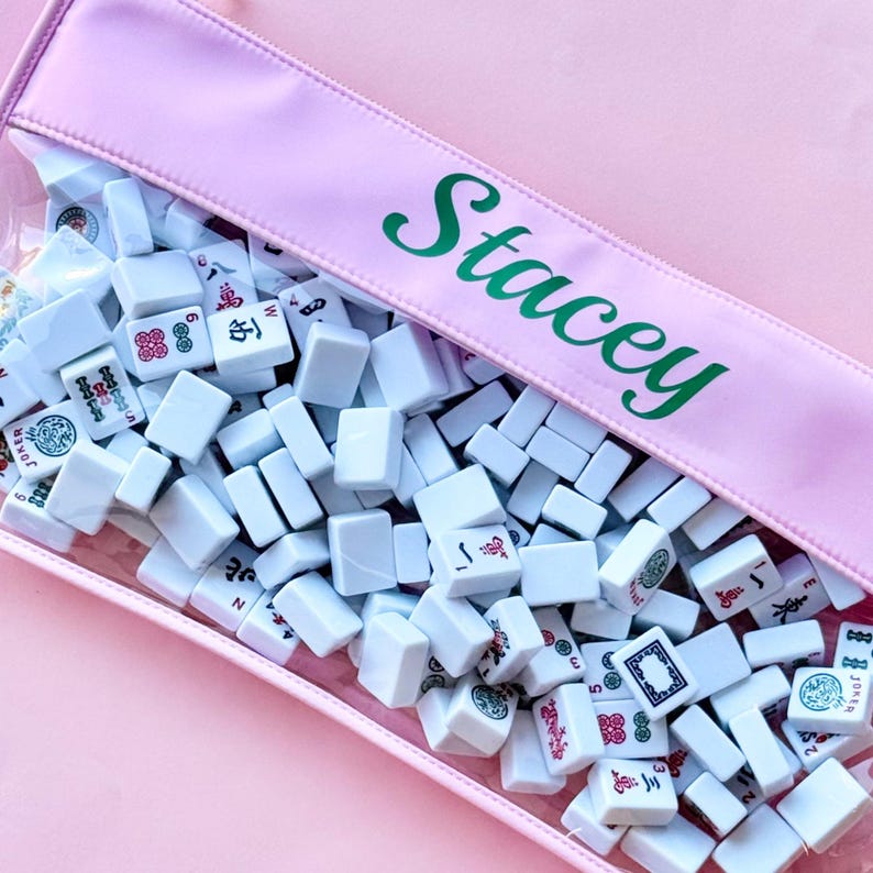 May include: A pink pouch containing white Mahjong tiles. The pouch has a clear front and a pink top with the name "Stacey" in green script. The tiles are white with various symbols and characters.