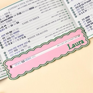May include: A pink and green decorative card with the name "Laura" printed in green. The card has a scalloped edge and a slot for sliding. Text on the card reads "NEWS 123 NEWS 456".