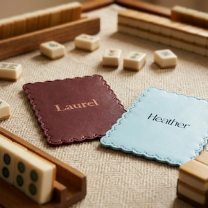 Könnte beinhalten: Nahaufnahme eines Mahjong-Sets mit Holzgestellen und Spielsteinen. Zwei Untersetzer mit gewelltem Rand sind zu sehen, einer bordeauxfarben mit "Laurel" und der andere hellblau mit "Heather". Die Spielsteine sind cremefarben mit grünen und roten Markierungen.