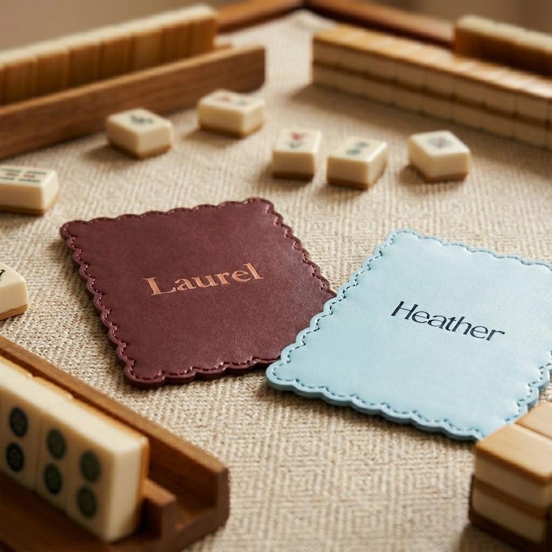 May include: A Mahjong set with wooden tile racks and cream-colored tiles with green and red markings. Two scalloped-edge coasters are in view, one burgundy with "Laurel" and the other light blue with "Heather" printed on them. The set is on a beige surface.