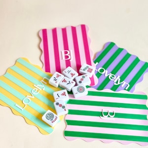 May include: A collection of white Mahjong tiles with red and green symbols, scattered on colorful, scalloped-edge mats. The mats feature vertical stripes in pink, green, yellow, and purple, with the text "Lovelyn" and initials.
