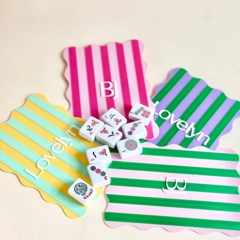 May include: A collection of white Mahjong tiles with red and green symbols, scattered on colorful, scalloped-edge mats. The mats feature vertical stripes in pink, green, yellow, and purple, with the text "Lovelyn" and initials.