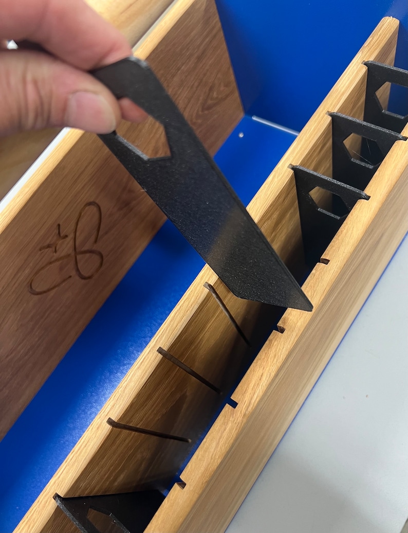 Pode incluir: Um organizador de ferramentas de madeira com uma tampa azul e v&aacute;rias ranhuras para ferramentas. Uma ferramenta preta com um furo hexagonal est&aacute; sendo colocada em uma das ranhuras.