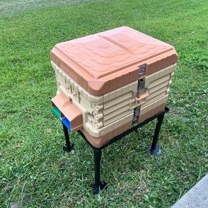 May include: A beige and brown plastic beehive with a black metal stand. The beehive has a hinged lid and a small opening on the side. The stand has four legs.