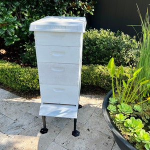 May include: A white wooden beehive with four stacked boxes and a white lid. The hive is elevated on a metal stand with black feet. The hive is outdoors, surrounded by greenery and a small pond.