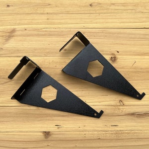 May include: Two black metal shelf brackets with a hexagonal cutout. The brackets are angled and have a small hook on the top.