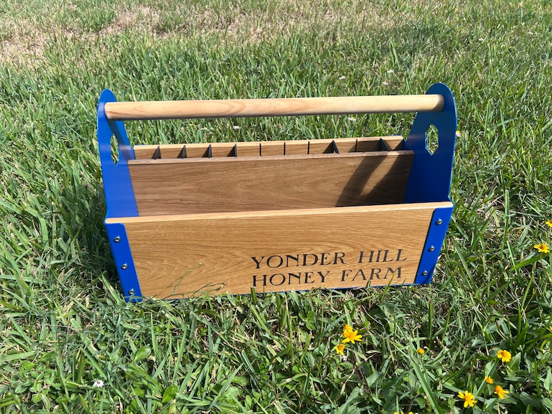 Pode incluir: Uma caixa de ferramentas de madeira com uma moldura de metal azul e uma al&ccedil;a de madeira. A caixa tem um r&oacute;tulo que diz "Yonder Hill Honey Farm".