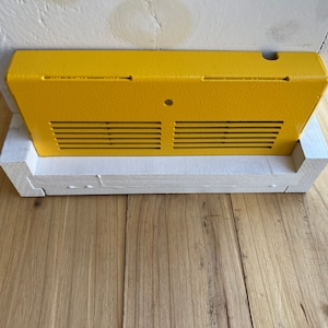 Op de afbeelding: Een gele metalen bijenkorf-ingangverkleiner met een ventilatieopening en een klein gaatje. De verkleiner is bevestigd aan een witte houten bijenkorf.