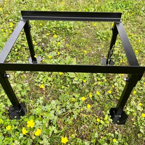 May include: Black metal stand with four legs and a rectangular top. The stand is sitting on a bed of green grass with yellow flowers.