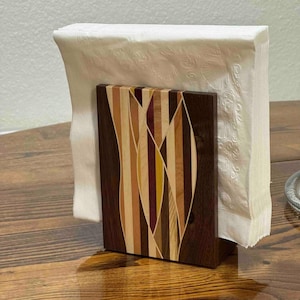 May include: A vertical wooden napkin holder featuring a central inlay of multi-colored wood strips in shades of dark brown, light tan, yellow, and deep purple, holding a stack of white paper napkins with a subtle embossed shell pattern.