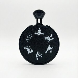 May include: A black, circular novelty item with a handle. The word "ASS" is printed on the item. White stick figures illustrate various sexual positions around the circle.