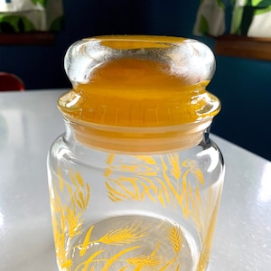 May include: A clear glass jar with a yellow lid and a wheat design. The jar is empty and has a yellow lid.