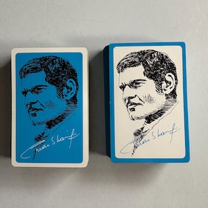 May include: Two decks of playing cards with a blue border and a portrait of a man in black ink. The man's portrait is on a white background. The signature "Omar Sharif" is written in cursive below the portrait.
