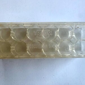 May include: A clear plastic thread box with the text "TRAUM" and "THREAD BOX" printed on the front. The box has 12 compartments for storing thread.