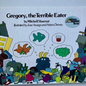 May include: A book cover with a white background and a cartoon illustration of a goat with a thought bubble above its head. The thought bubble contains a green fish, a glass of orange juice, and a bunch of green broccoli. The book title is "Gregory, the Terrible Eater" by Mitchell Sharmat, illustrated by Jose Aruego and Ariane Dewey. The book is part of the Reading Rainbow Book series.