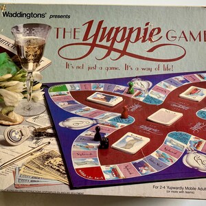 May include: The Yuppie Game board game box features a red and blue game board with a white game piece and a glass of wine. The box is decorated with a white flower and a gold-colored pen. The text on the box reads "The Yuppie Game" and "It's not just a game. It's a way of life!"