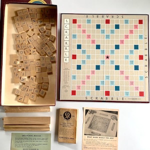 May include: A vintage Scrabble game set with wooden tiles, a game board, and instructions. The game board has a red and blue design with a star in the center. The tiles are in a wooden box with a red lid. The instructions are on a white card with a red and blue logo.