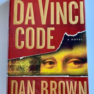 May include: A red hardcover book with the title "The Da Vinci Code" in gold lettering. The author, Dan Brown, is listed below the title, along with the text "A Novel" and "Author of Angels & Demons". The cover features a close-up of a woman's eyes from the Mona Lisa painting.