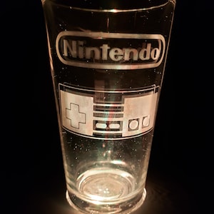 May include: A clear glass pint glass with an etched design of the Nintendo logo and a controller. The glass is illuminated from below, casting a warm glow.