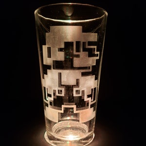 May include: A clear glass pint glass with a white etched design of a pixelated video game character. The glass is sitting on a black surface.