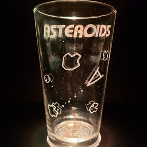 May include: A clear glass with the word "ASTEROIDS" etched on the side. The glass is decorated with a spaceship and asteroids.