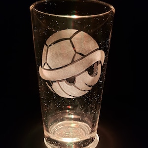 May include: A clear glass pint glass with a white etched design of a Koopa Troopa shell from the Super Mario Bros. video game series.
