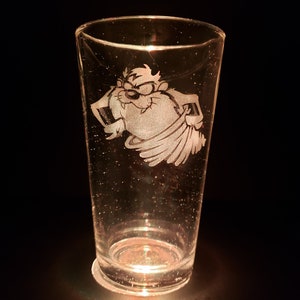 May include: A clear glass with a white etched design of the cartoon character Taz the Tasmanian Devil. The character is depicted in a swirling motion.
