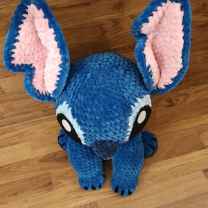 May include: A plush blue creature with large, pink-lined ears and black eyes. The handmade toy has a textured, fuzzy appearance and is posed sitting upright. The creature's feet and nose are also blue, with black claws. A charming, soft toy.
