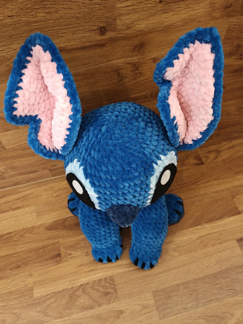 May include: A plush blue creature with large, pink-lined ears and black eyes. The handmade toy has a textured, fuzzy appearance and is posed sitting upright. The creature's feet and nose are also blue, with black claws. A charming, soft toy.