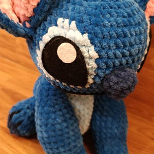 May include: A handmade blue plush toy with large white-rimmed eyes, black accents, and pink ears. The plushie is made of a soft, textured material and has a friendly, cartoonish appearance. The toy is sitting on a wooden surface.