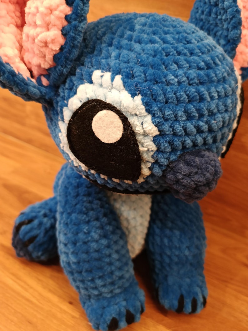 May include: A handmade blue plush toy with large white-rimmed eyes, black accents, and pink ears. The plushie is made of a soft, textured material and has a friendly, cartoonish appearance. The toy is sitting on a wooden surface.