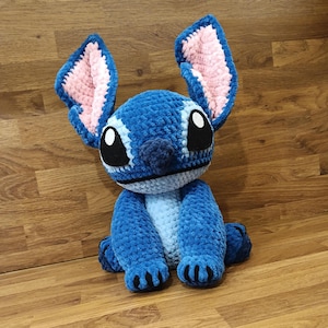 May include: A blue crochet plush toy with large ears, black eyes, and a light blue belly. The ears have pink inner linings. The toy is sitting on a wooden surface. The plush toy is a handmade item.