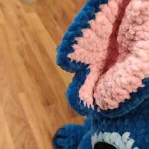 May include: A close-up of a blue and pink plush toy, featuring a large ear with a blue border and pink interior. The toy has a black and white eye and a blue body. The plush is made of a soft, textured material.
